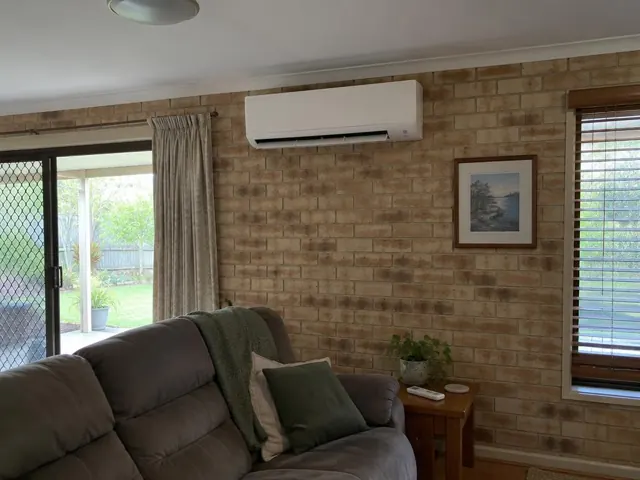 Clean indoor split system on a brick living room wall with louvre open, beside a sofa and windows in Coombabah